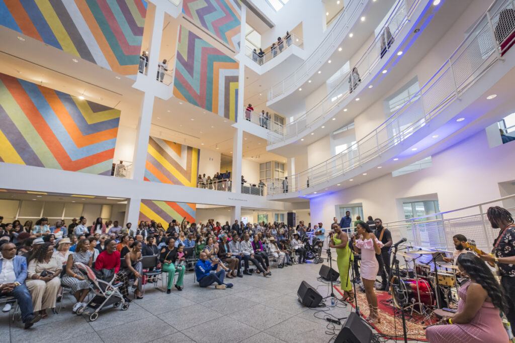 A live band performs for a large, seated audience in a modern, brightly lit atrium with colorful geometric wall art and multiple balcony levels filled with people.